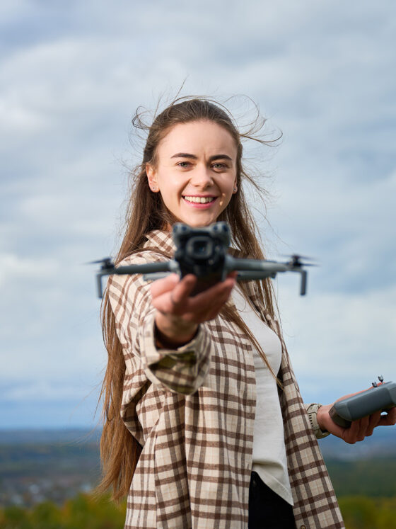 A woman, smiling, holding a mini sized DJI drone in her hand, pointing it forward to the camera. In her other hand she holds the controller for the drone.