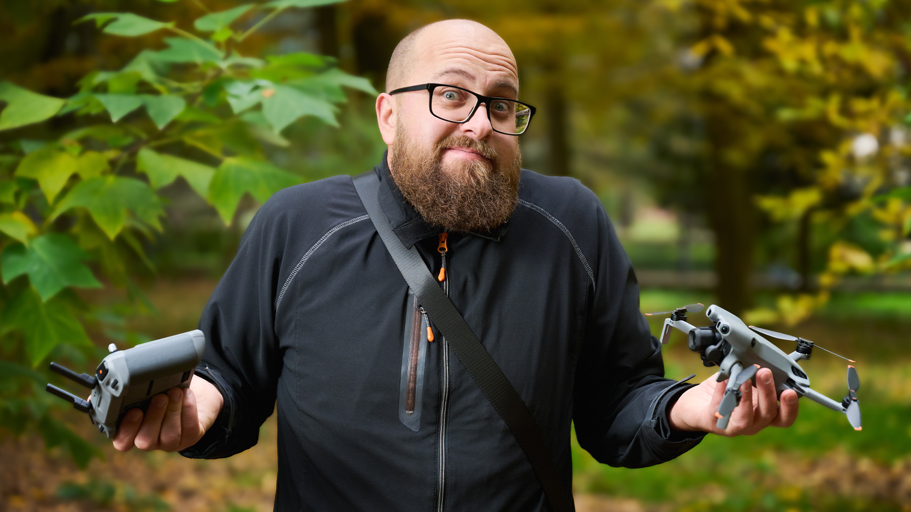 A male person, with glasses and beard, holding a DJI mini drone and controller in their hands. He is shrugging and making a face expression which shows us he is not sure about what to do.