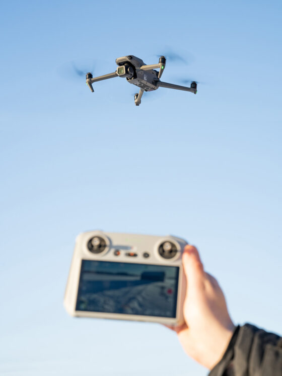 Medium-sized DJI drone in the air, clean blue sky in the background. A hand holding the RC controller.